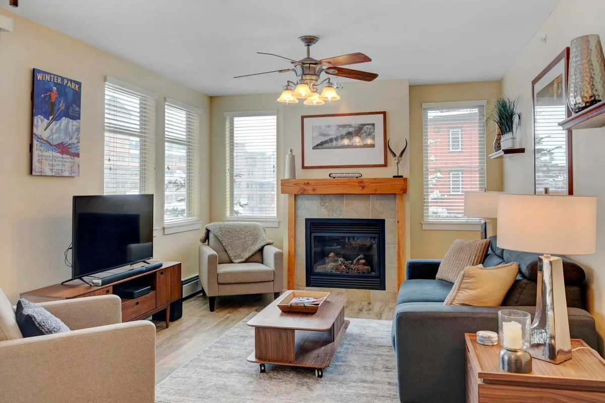 A cozy living room featuring a rustic stone fireplace as the focal point, surrounded by a plush sofa and coffee table. Artwork adorns the walls, and large windows let in ample natural light, creating a warm and inviting space. The arrangement encourages socializing or relaxing by the fire.