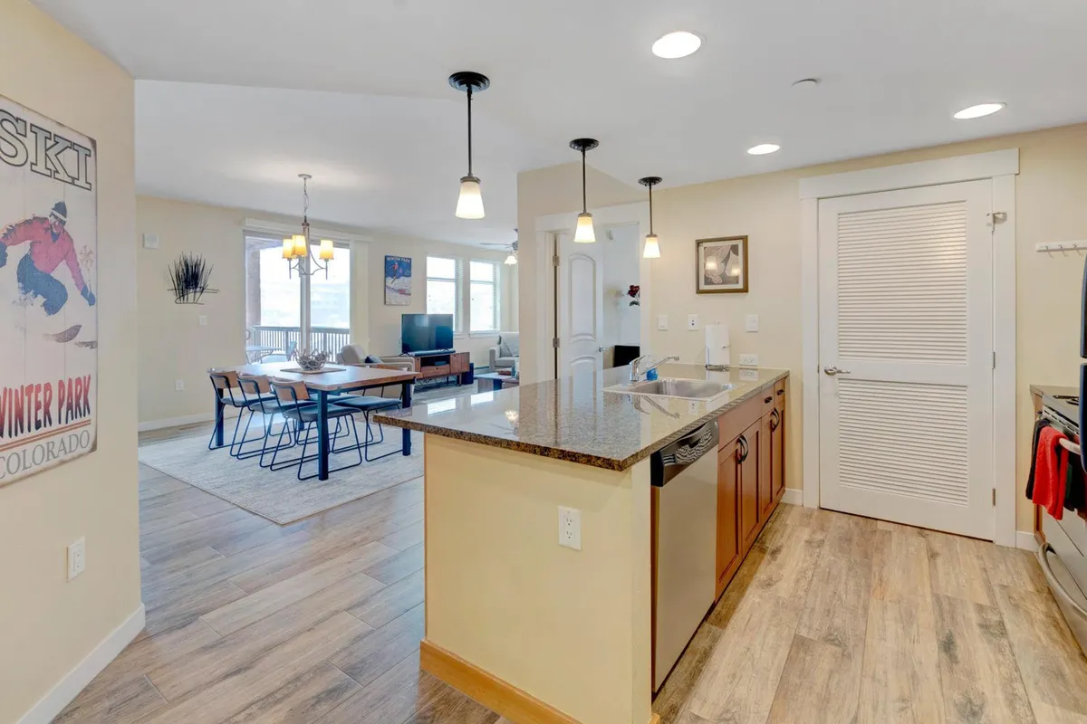 An open-concept living space that seamlessly connects the living room, dining area, and kitchen. Granite countertops, a central kitchen island, and a well-appointed dining area are visible, with large windows framing the serene mountain scenery. The space is filled with warm tones and high-quality furnishings.