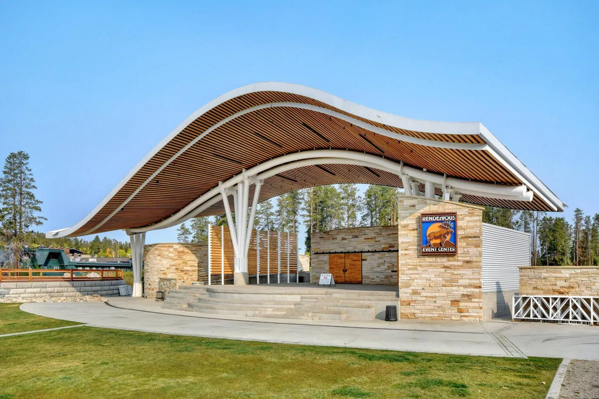 An exterior shot featuring a curved roof structure and sturdy stone walls, with visible event signage in the foreground. The scene is set for an outdoor event or concert, and groups of people are likely gathering around, enjoying the lively atmosphere and the beautiful mountain setting.