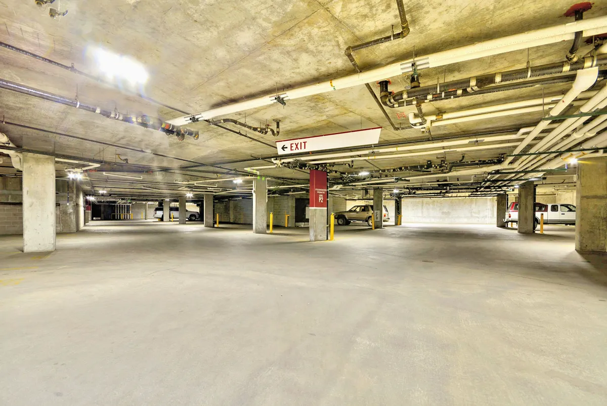 An underground parking garage with visible parking spaces, vehicles, support columns, and an exit sign. The space is clean, well-lit, and secure, emphasizing the convenience and safety of heated underground parking for residents and guests.