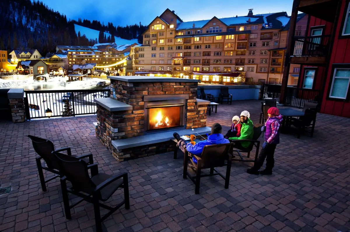 An inviting outdoor plaza set against a snowy mountain backdrop, featuring a large circular hot tub surrounded by comfortable seating, a double-sided gas fireplace, and illuminated buildings. The scene captures guests gathered around the fire, enjoying warm drinks, and soaking in the hot tub under twinkling lights, conveying a sense of warmth, togetherness, and après-ski relaxation.