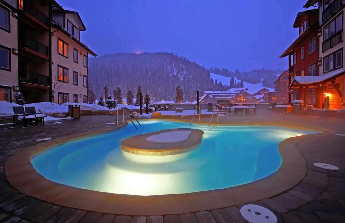 A heated outdoor pool set against a snowy landscape with softly illuminated buildings, radiating a tranquil and inviting atmosphere perfect for relaxing after an adventurous day on the mountain.