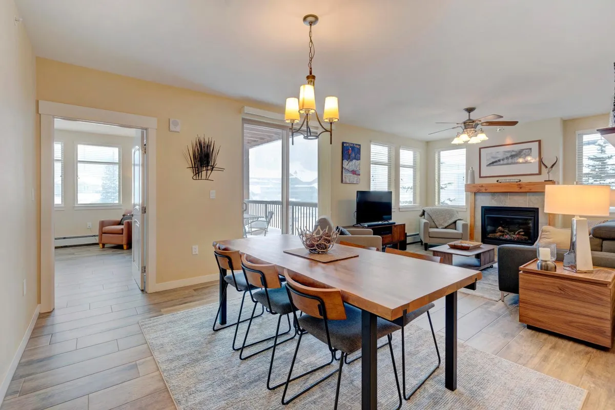 A welcoming dining area adjacent to the living space, featuring a dining table set near a fireplace and large windows. Comfortable seating and mountain-inspired decor enhance the inviting atmosphere, while the windows offer glimpses of the outdoors.
