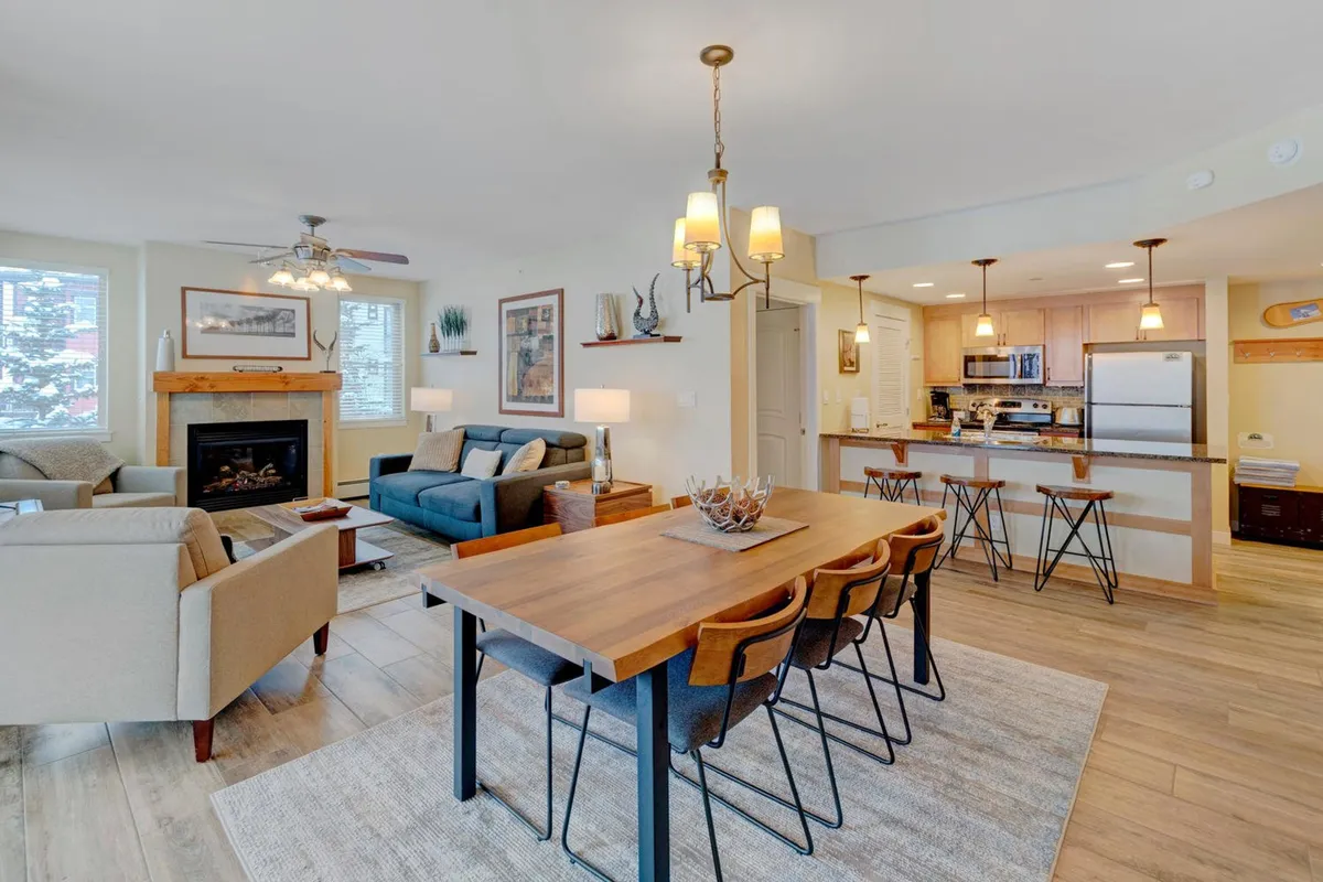 The hero image showcases a warm and inviting open-plan living area, featuring a crackling fireplace, a comfortable sofa, a stylish dining table set for gatherings, and a modern kitchen. The scene emanates a cozy, welcoming atmosphere perfect for relaxing, socializing, and enjoying meals together after a day on the slopes.