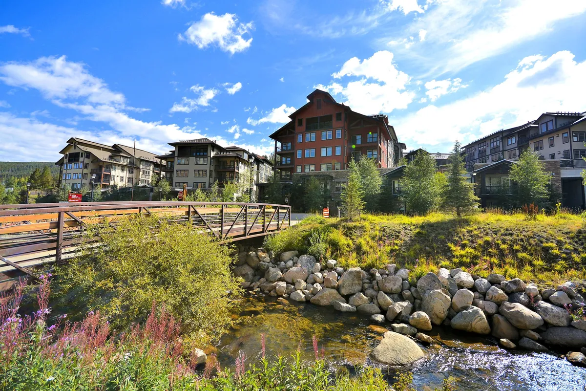 A tranquil wooden bridge spans a meandering stream, framed by lush greenery and surrounded by nearby buildings. The scene is bathed in natural light, creating a peaceful, community-oriented vibe that invites exploration and relaxation in nature.