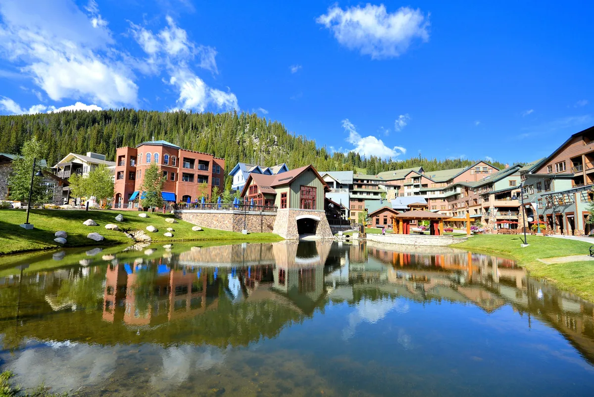 A tranquil pond reflects the colorful buildings and a charming bridge, creating a cheerful yet serene environment. The setting highlights outdoor gathering spaces and scenic strolls, suggesting peaceful moments and opportunities for exploration just outside the retreat.