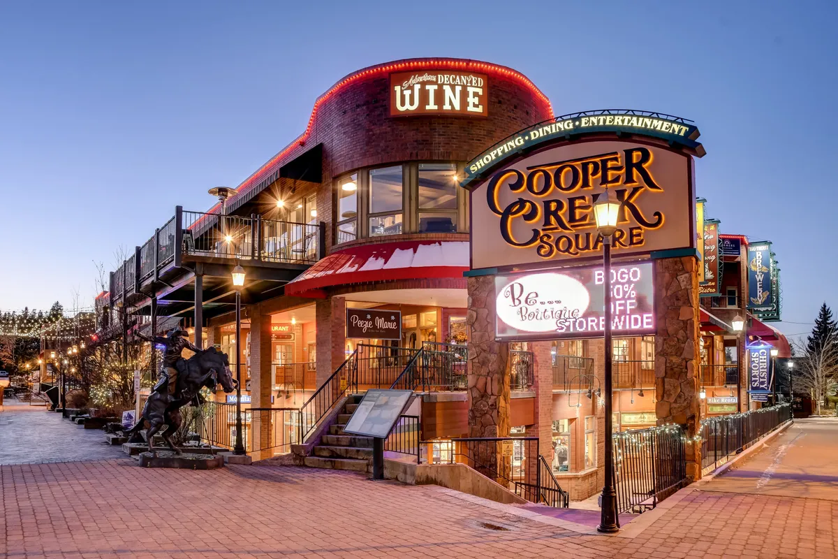 The vibrant scene at Cooper Creek Square features shop signs, a wine shop, outdoor seating, and a distinctive horse sculpture. People can be seen dining, shopping, and socializing, with festive touches that hint at lively village life just steps from the retreat.