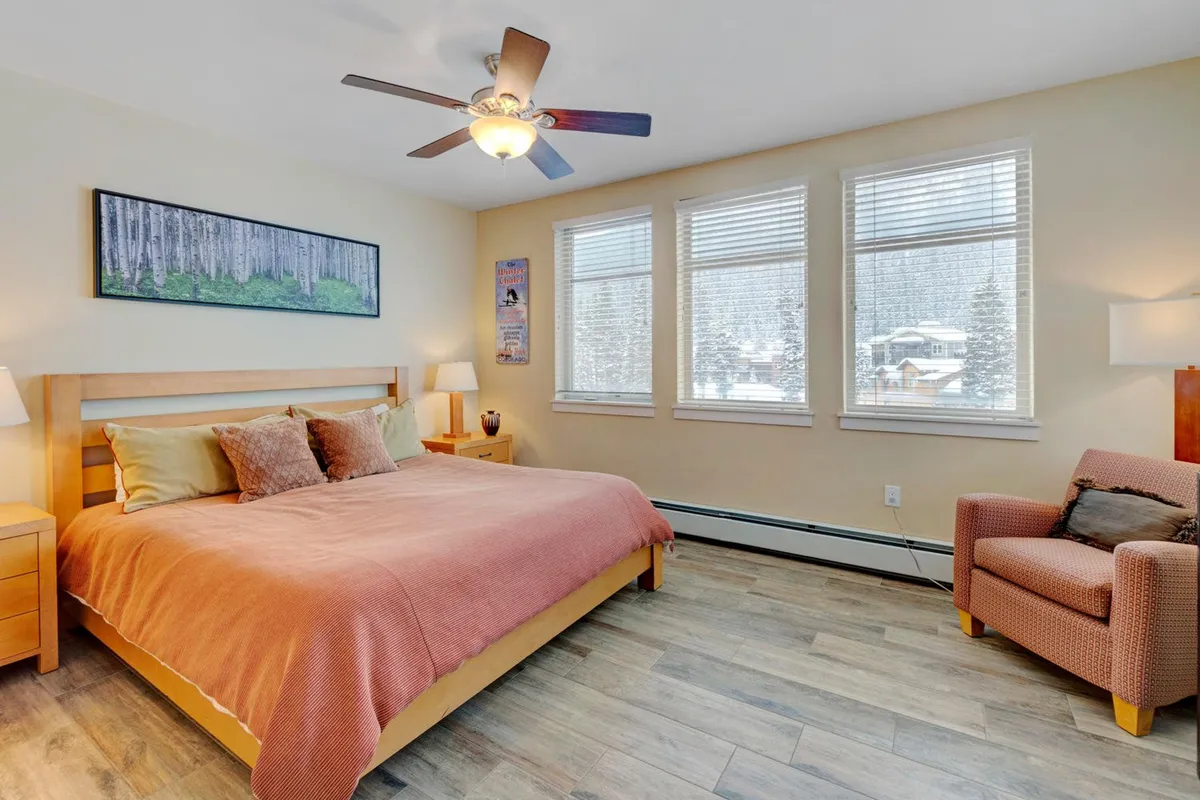 A serene bedroom featuring a plush bed with crisp linens, soft artwork above the headboard, large windows allowing natural light to pour in, and a comfortable armchair situated for enjoying tranquil views.