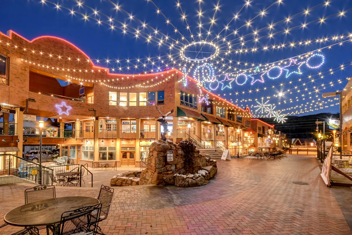 An exterior view of a lively plaza with string lights, decorative stars, a central fountain, and outdoor seating. The building facade and festive decorations create a joyful atmosphere, perfect for dining, shopping, and socializing during local festivals or holiday events.